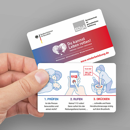 Eine Hand hält zwei Info-Scheckkarten (Forder- und Rückseite) mit den abgebildeten Maßnahmen, welche im Falle einer nötigen Wiederbelebung zu beachten sind. 